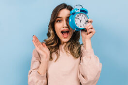 woman-with-wavy-hair-holding-clock-studio-shot-of-amazed-girl-isolated-on-blue-background
