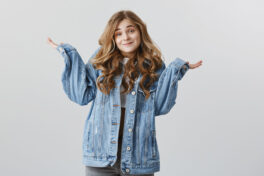 Who knew this could happen. Portrait of careless questioned attractive female coworker in stylish denim jacket, shrugging and smiling awkwardly, having no idea or being unaware over gray wall